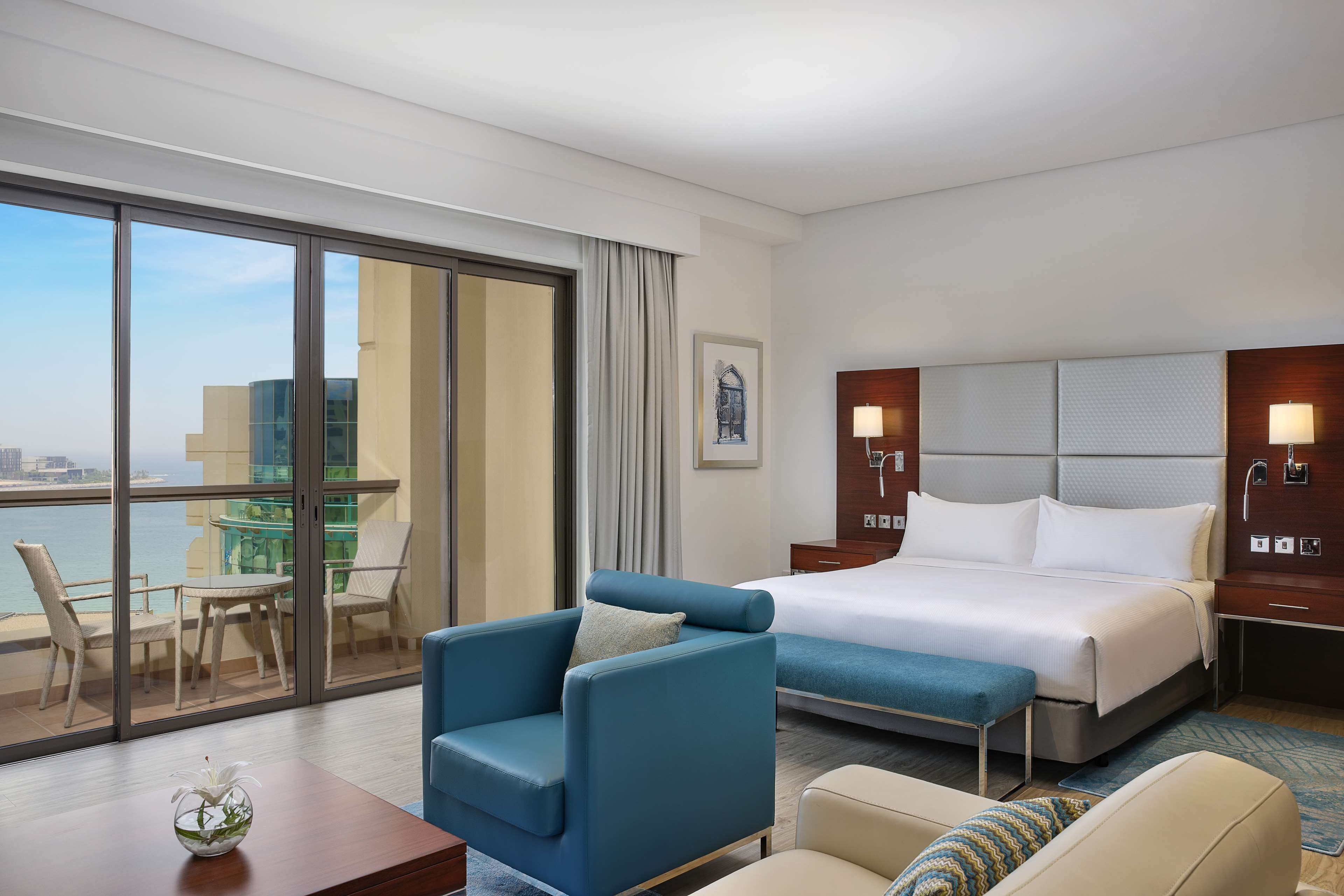 Habitación de hotel con una cama grande, zona de estar con una silla y una mesa, vista de la ciudad a través de amplios ventanales, y un balcón con mesa y sillas.