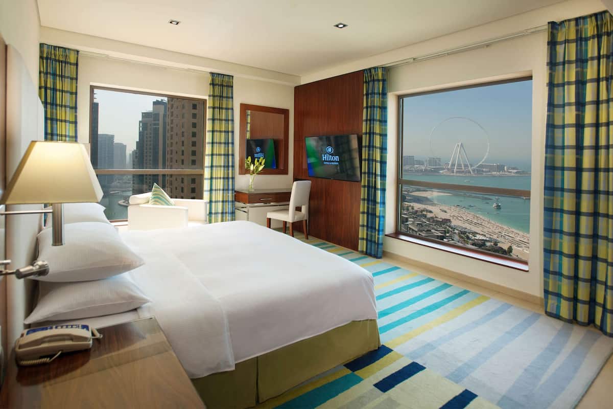 A hotel room with a bed, a desk with a lamp, a phone, a TV, and a large window with a view of a beach and a Ferris wheel.