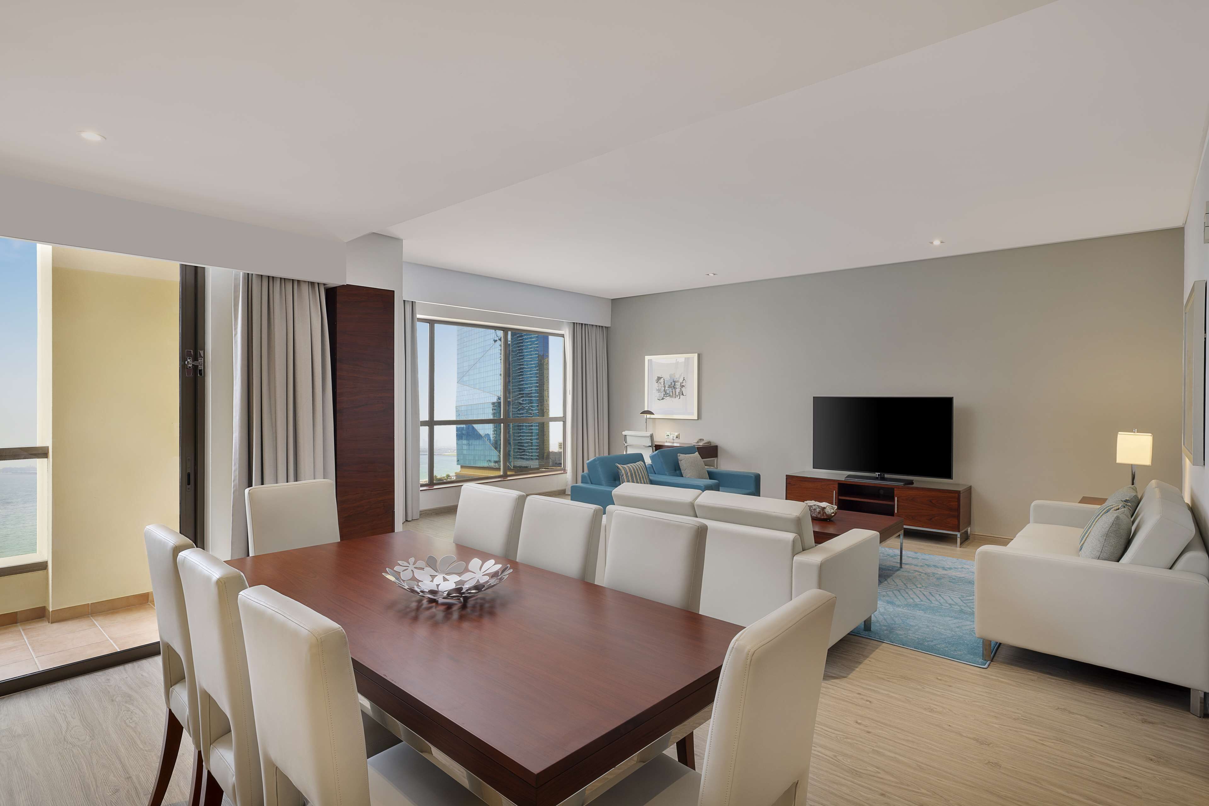 A modern living room with a dining area, a flat-screen TV, and a large window offering a city view.