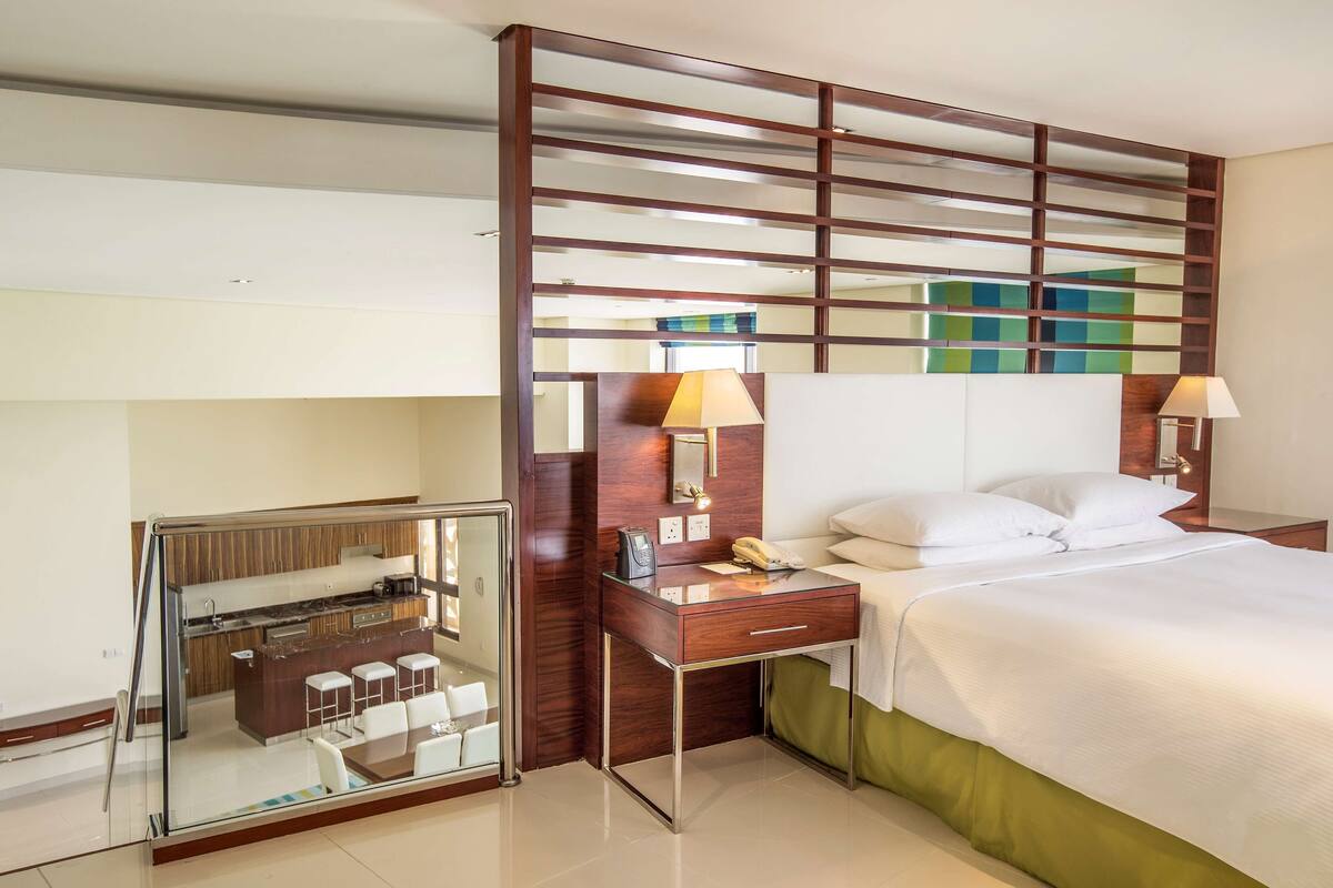 A hotel room with a bed, bedside lamps, a desk with a telephone, and a glass partition separating the room from a kitchenette area.