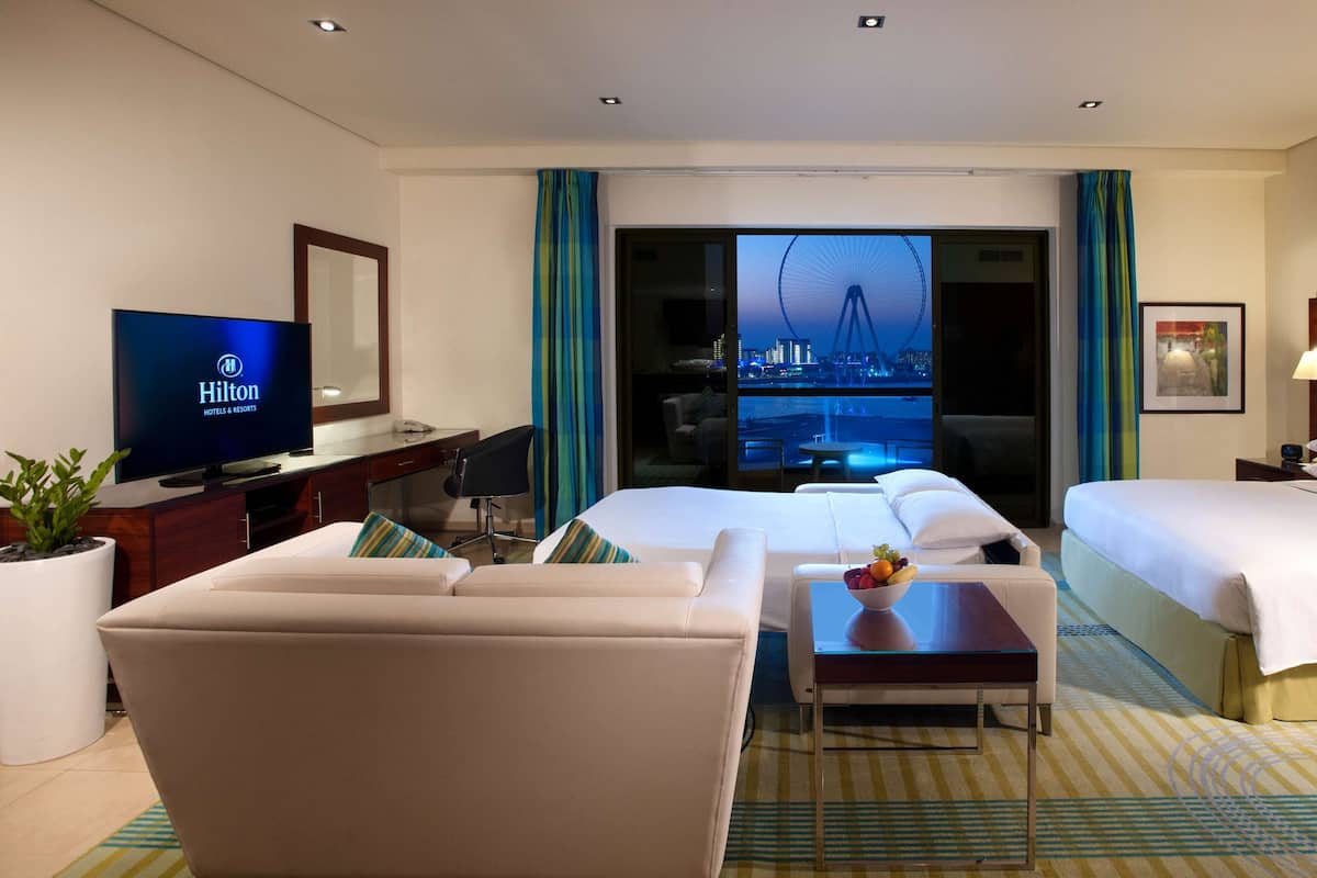 A hotel room with two beds, a TV, a desk, a chair, a small table with fruit, and a view of a cityscape.