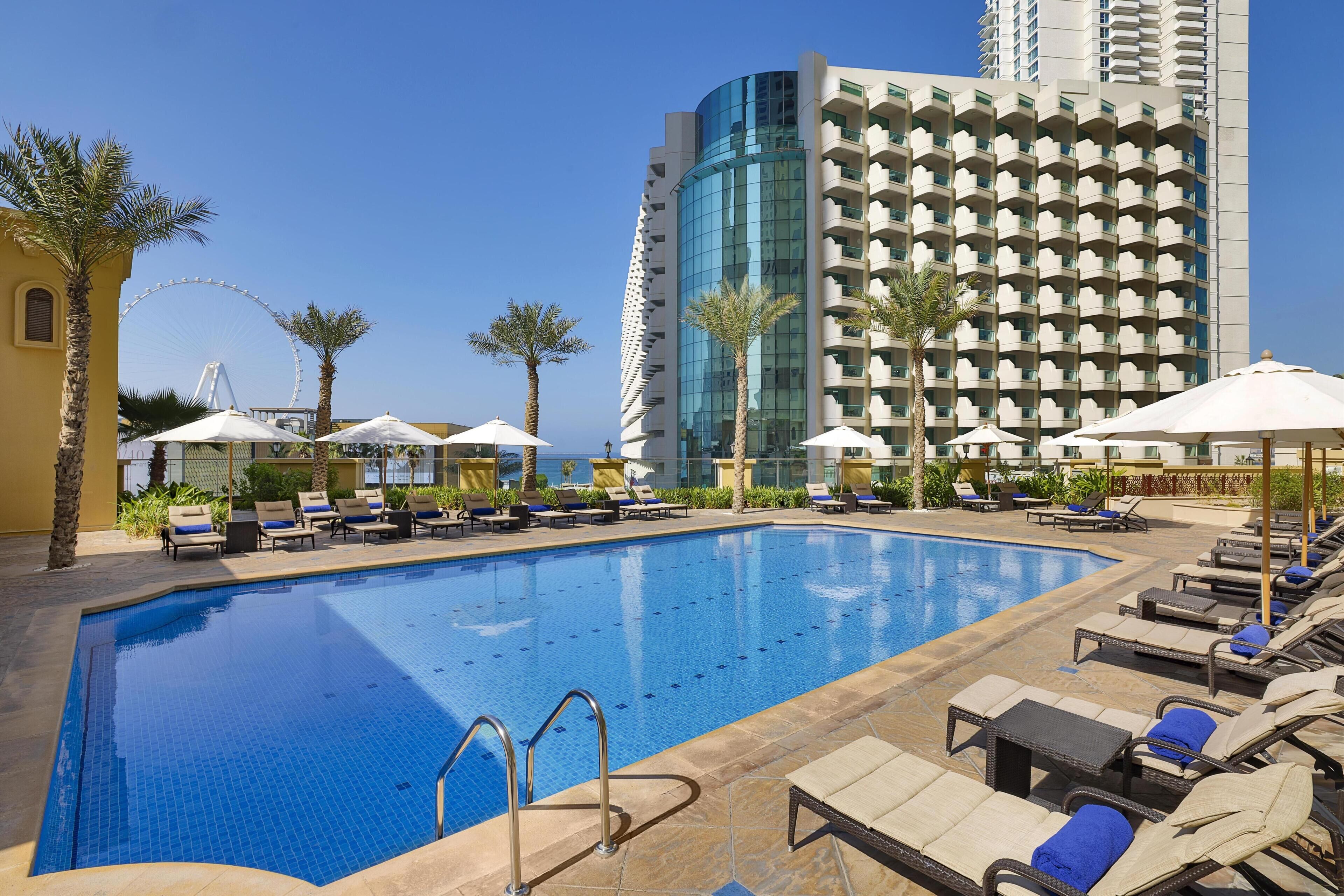 A hotel pool area with lounge chairs, umbrellas, and a tall building in the background.