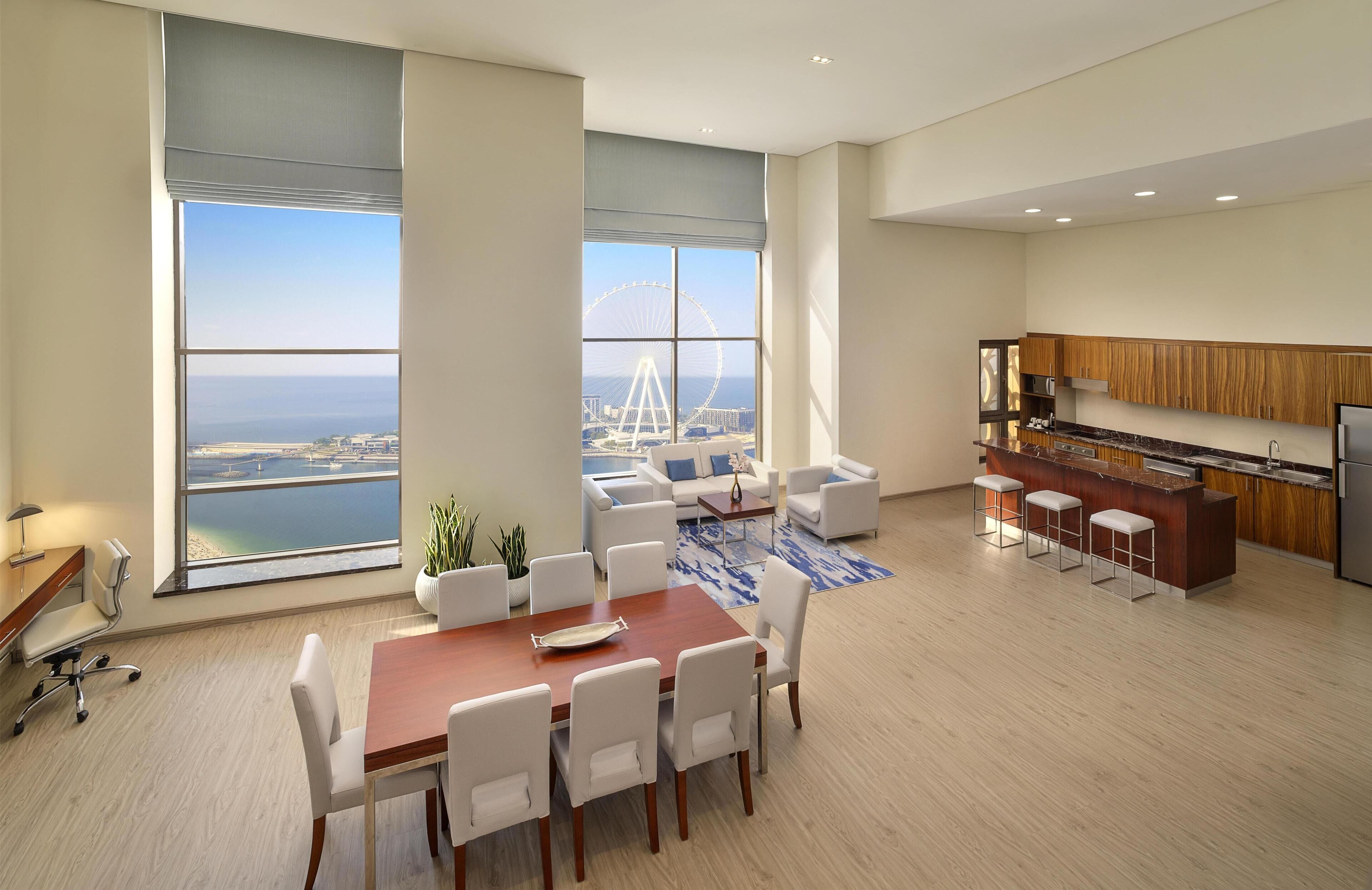 A modern living room with a dining area, large windows with a view of a Ferris wheel, and a kitchen with wooden cabinets.