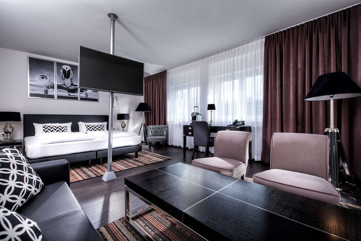 A modern hotel room with a bed, a desk, two chairs, a coffee table, and a TV.