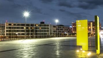 Un edificio moderno con varios pisos, una plaza pavimentada con un letrero amarillo de indicaciones y farolas.