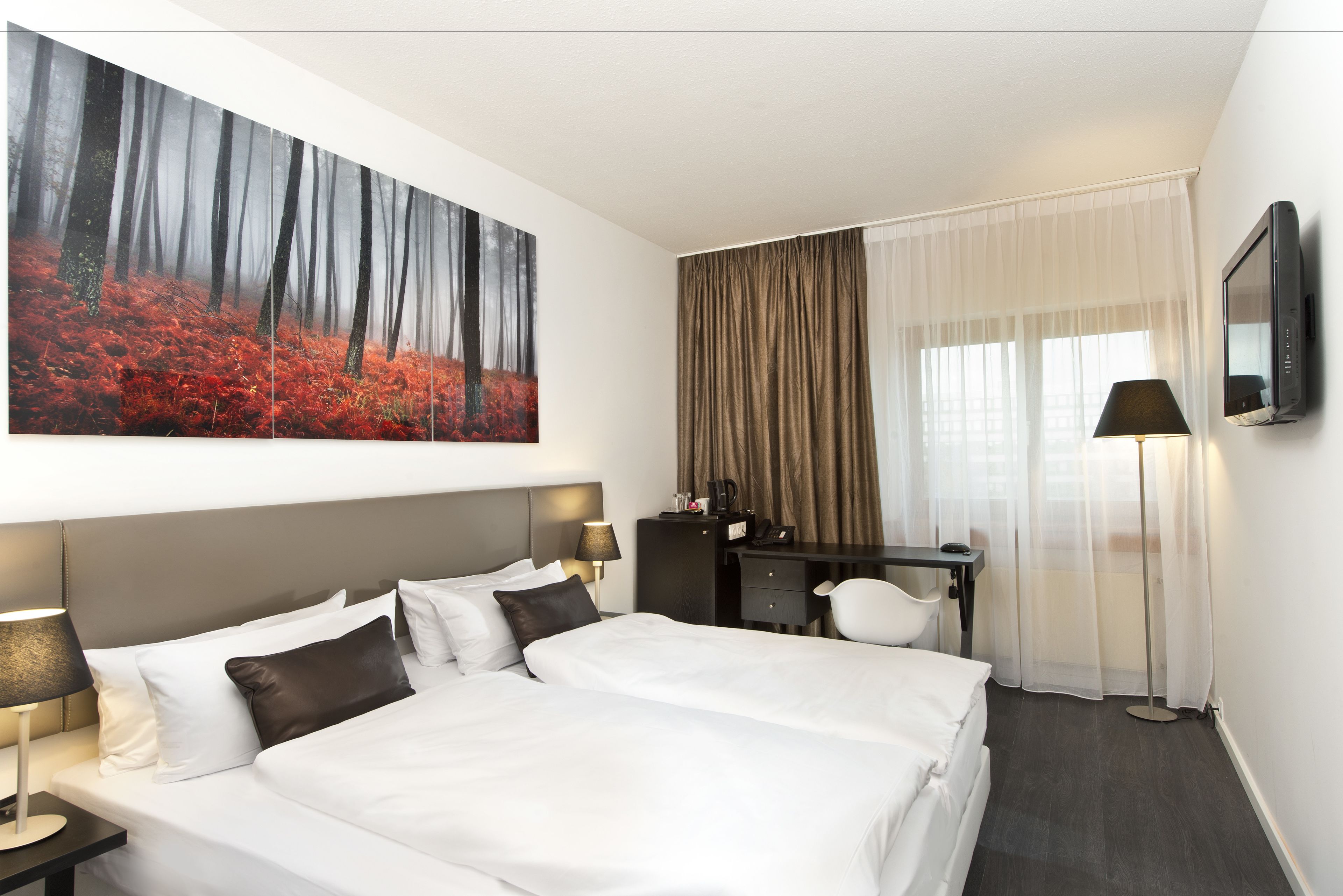 A hotel room with a large bed, a desk, a chair, a flat-screen TV, and a painting of a forest.
