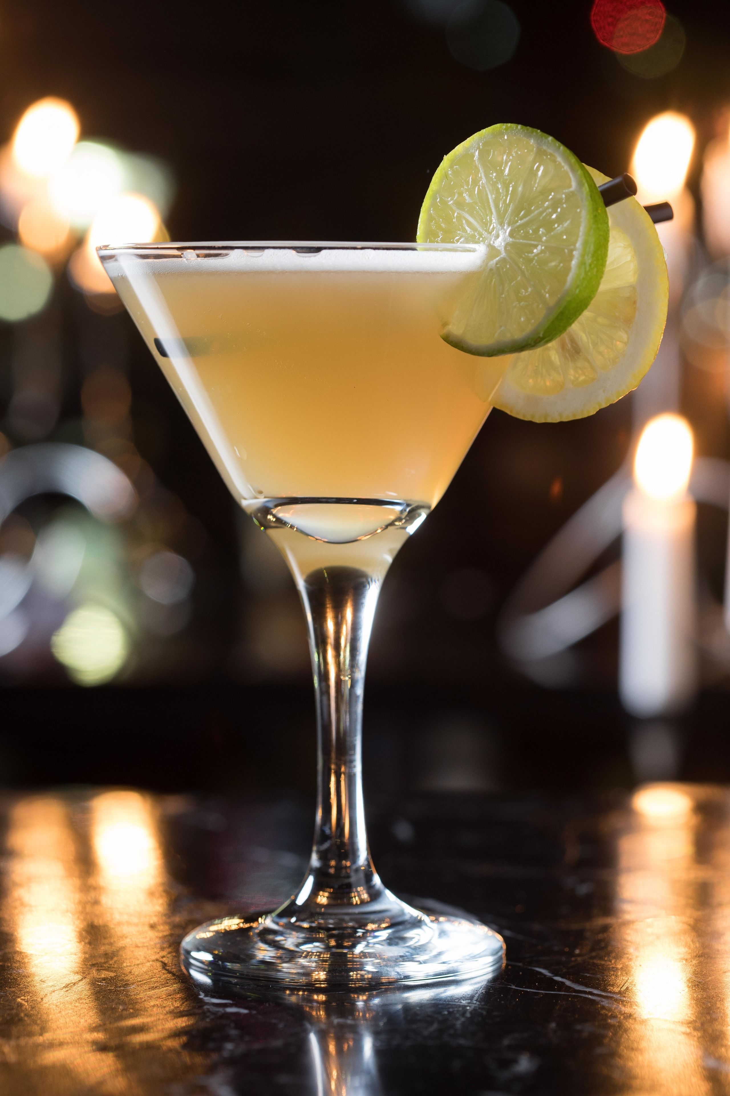 Un cocktail servi dans un verre à martini, garni d’une tranche de citron vert, posé sur une surface réfléchissante.