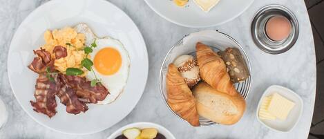 A breakfast setting with plates of eggs, bacon, croissants, and fruit.