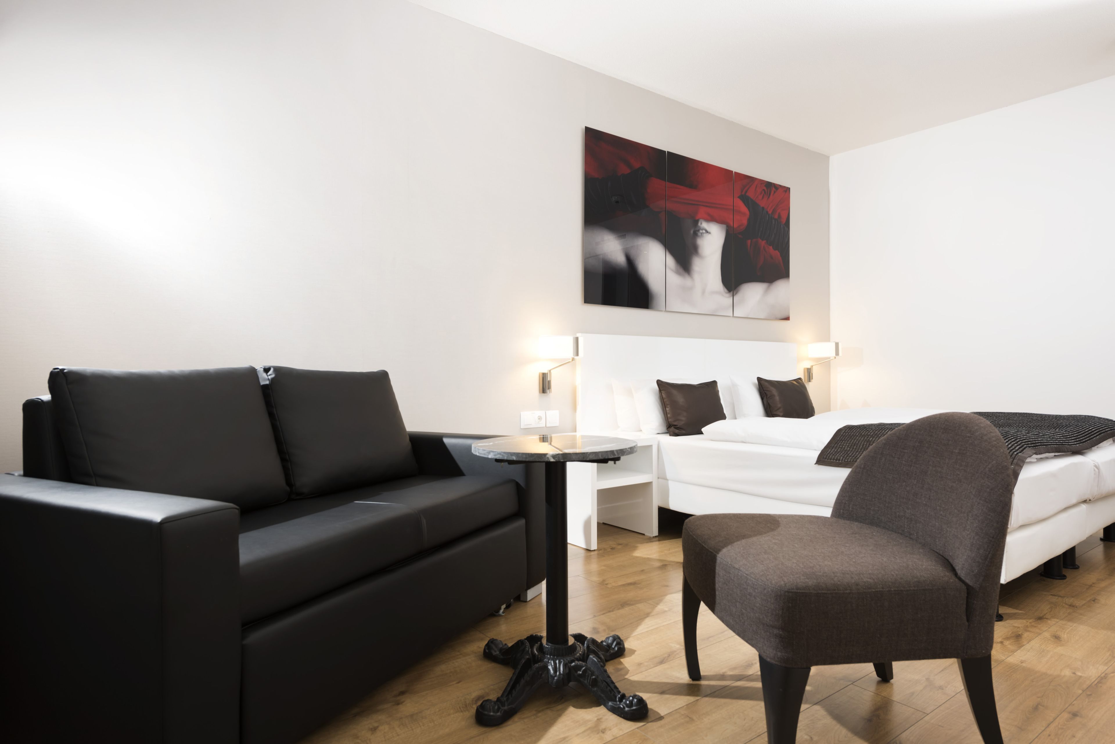 Una habitación de hotel moderna con un sofá negro, una mesa redonda, un sillón gris, una cama blanca y un cuadro abstracto grande en la pared.