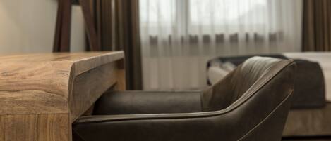 A modern hotel room with a wooden desk, a leather armchair, and a large window with sheer curtains.