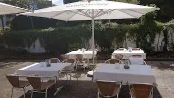 An outdoor dining area with white tables and wicker chairs, covered by large umbrellas with the Gerolsteiner logo.