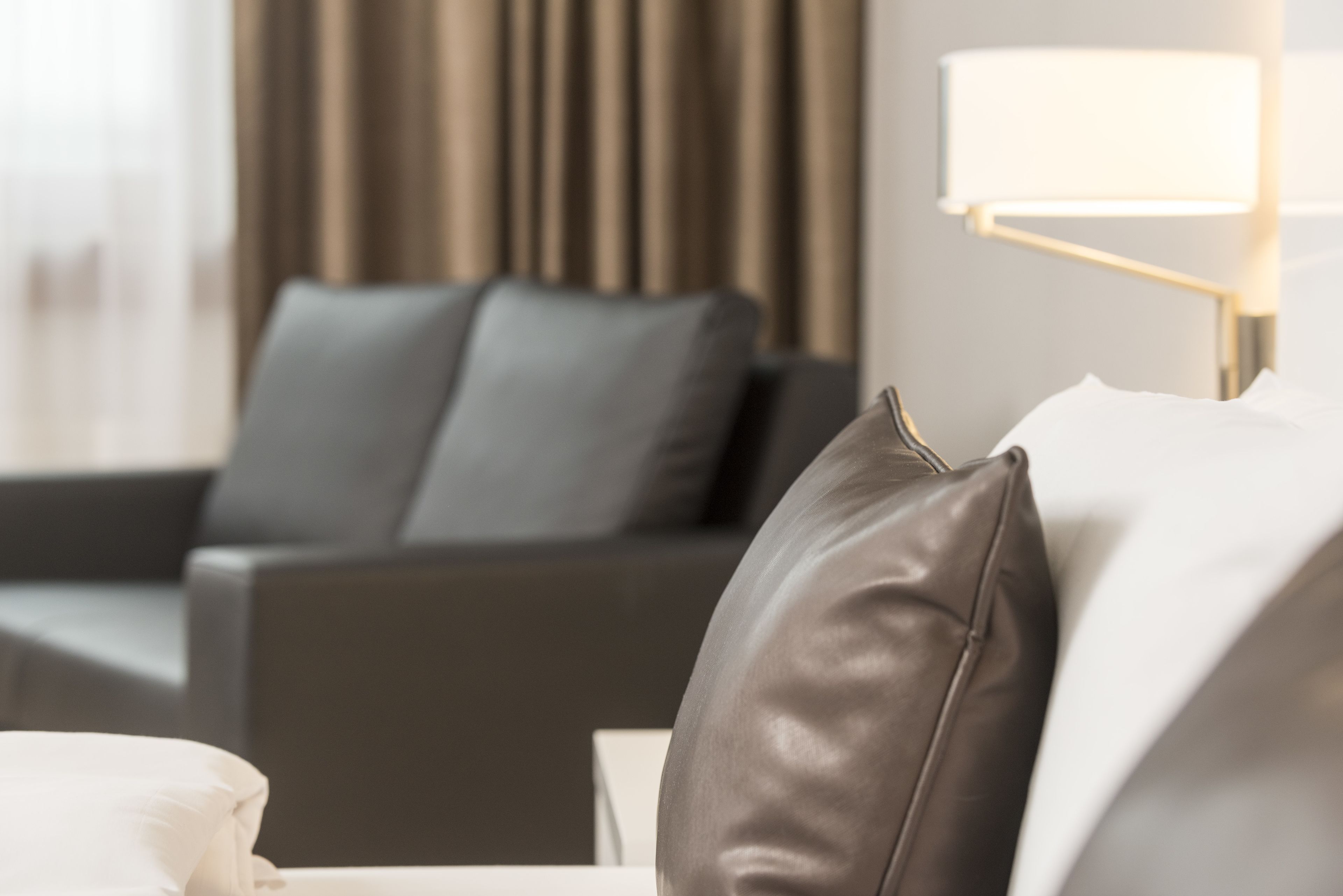 A modern hotel room with a grey sofa, a white pillow, and a bedside lamp.