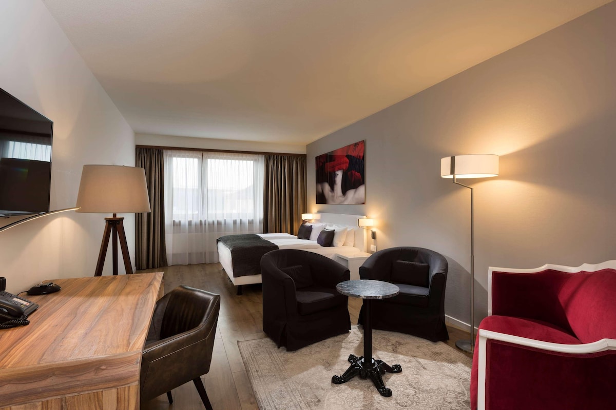 A hotel room with a bed, a desk with a telephone, two black armchairs, a red armchair, a small round table, and a floor lamp.