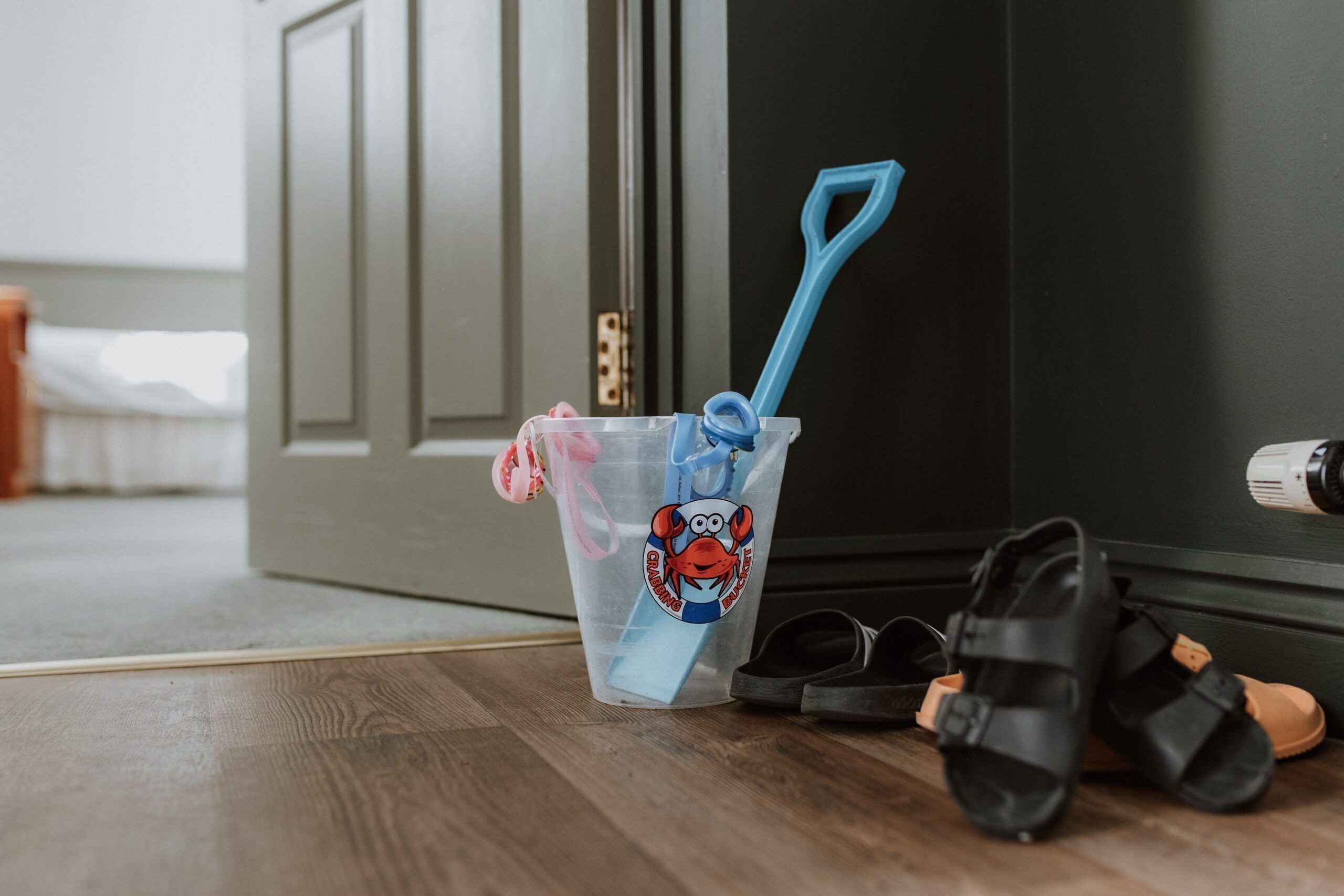 Un vaso de plástico con diseño de cangrejo, una pala azul y un par de sandalias sobre un piso de madera.