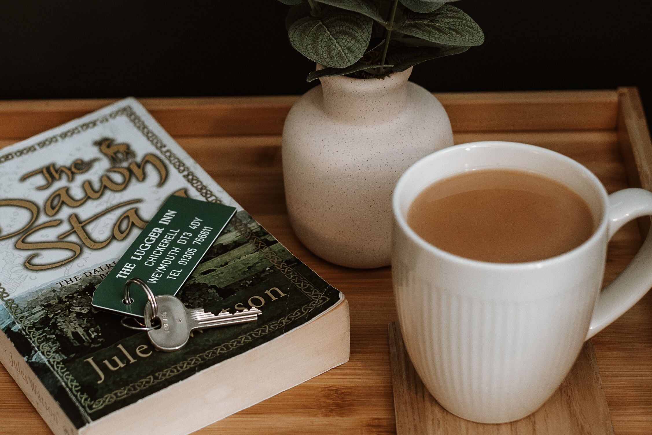A book titled 'The Dawning' with a keychain on top, a white mug with a beverage, and a vase with green leaves on a wooden tray.