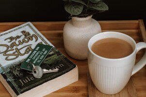 Un libro con el título 'The Dawning', un llavero encima, una taza blanca con una bebida y un florero con hojas verdes sobre una bandeja de madera.