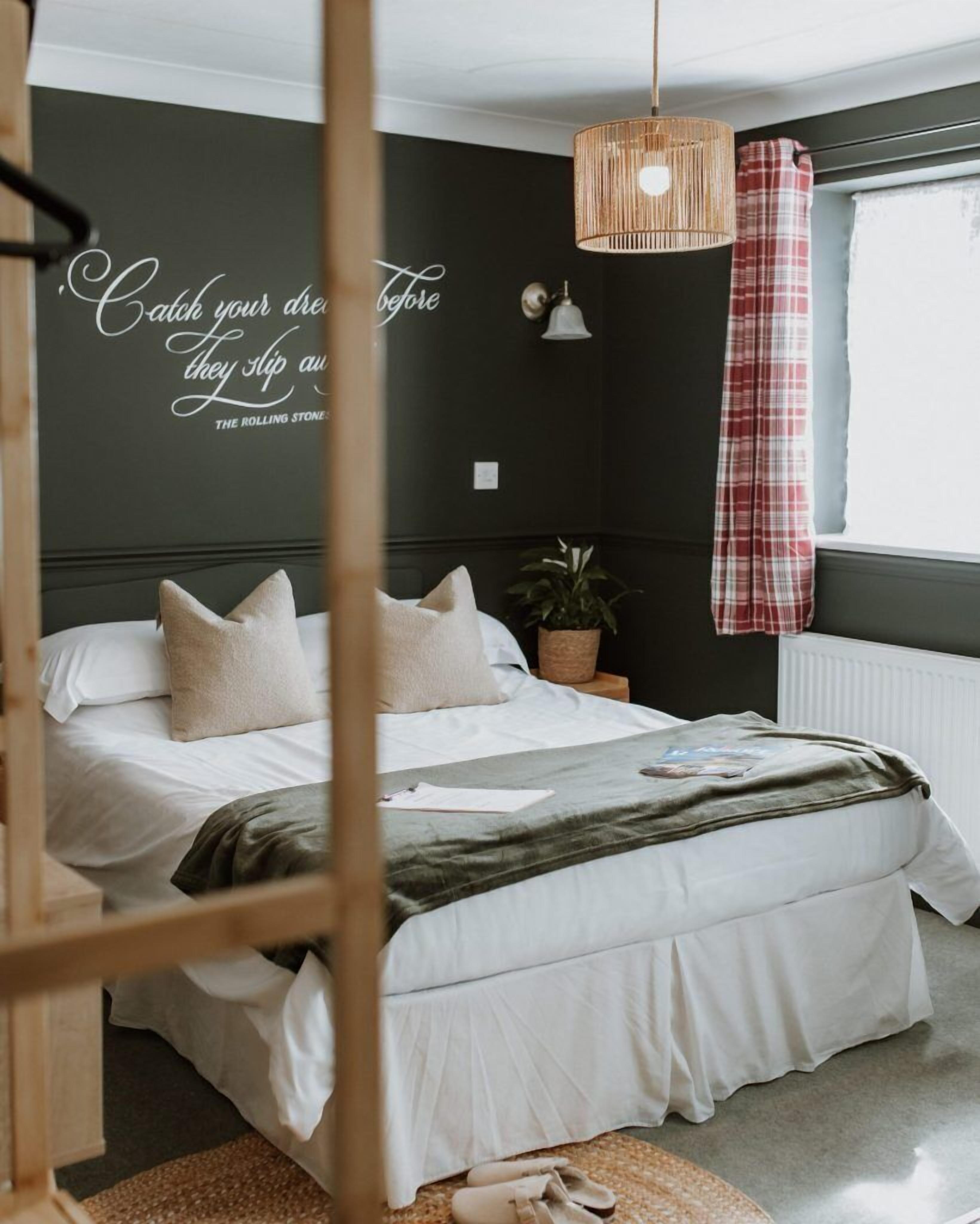 A bedroom with a bed, a wall with a quote, a hanging light, a window with a curtain, and a plant.