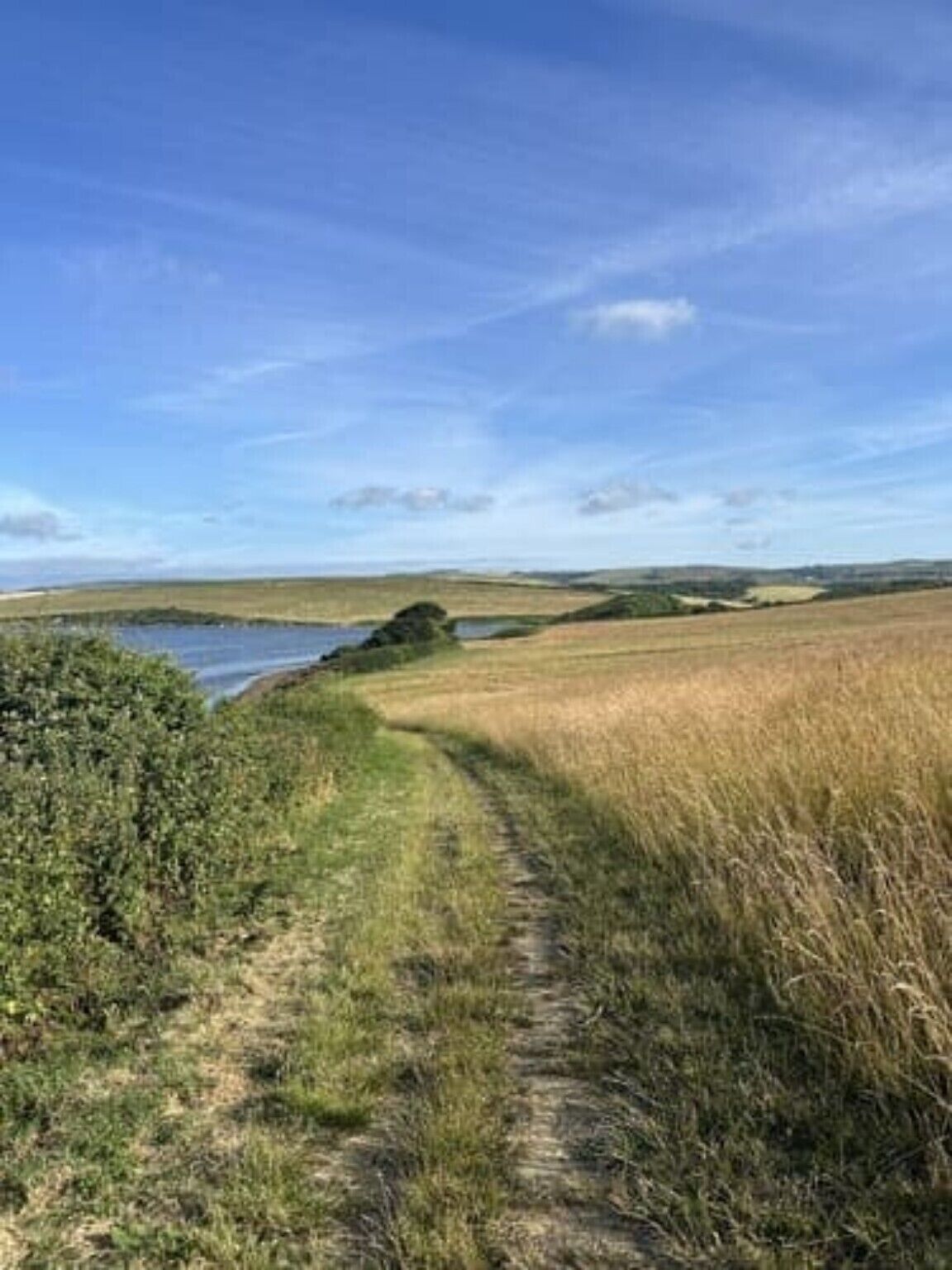 A rural landscape with a dirt path leading to a body of water, surrounded by fields and distant hills.