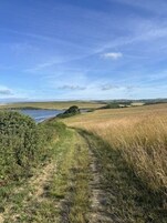 Un paisaje rural con un camino de tierra que conduce a una masa de agua, rodeado de campos y colinas al lejano.