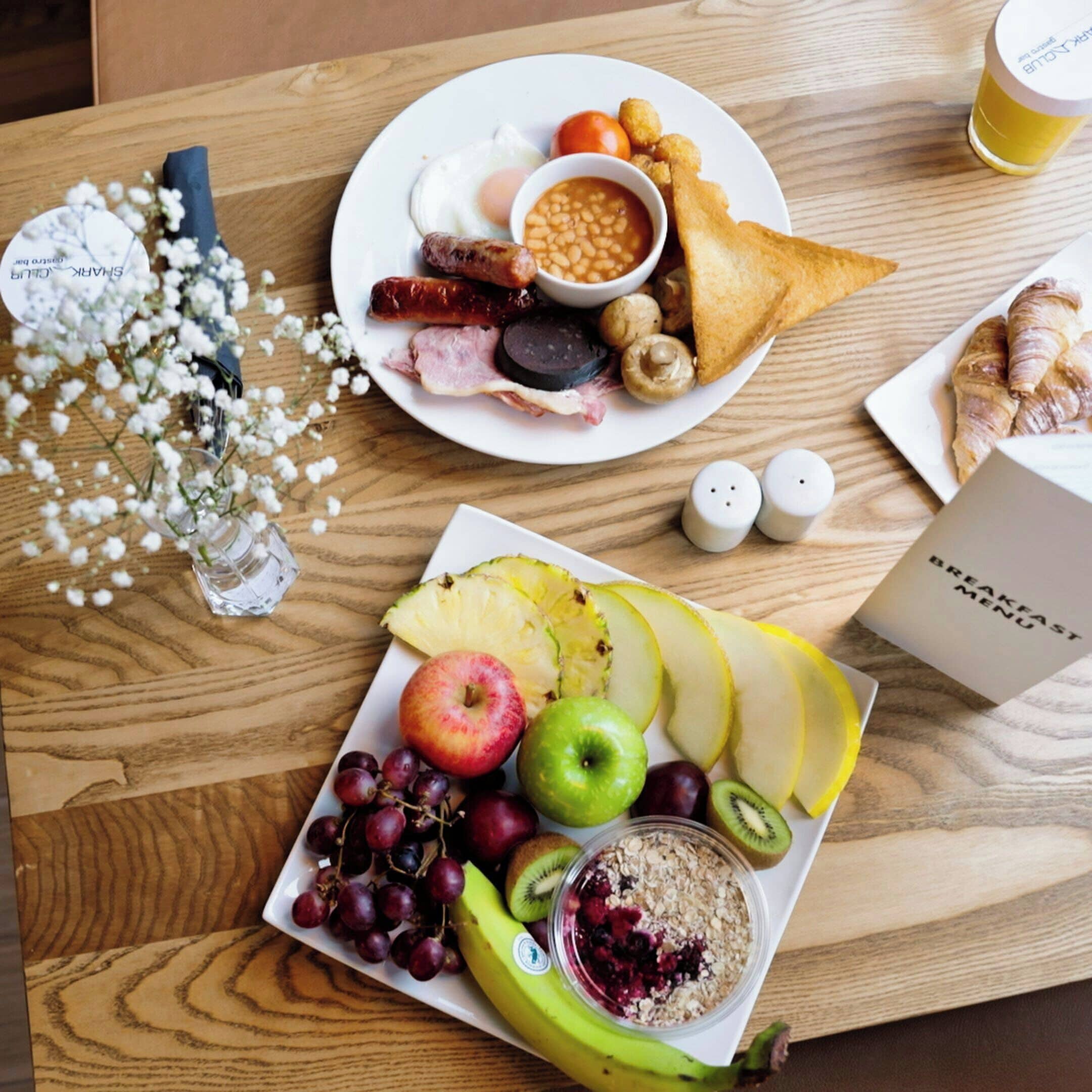 Ein Frühstücksteller mit Eiern, Würstchen, Bohnen, Pilzen und Toast, dazu ein Obstteller mit Äpfeln, Trauben, einem Kiwi und einer Banane mit Beeren.