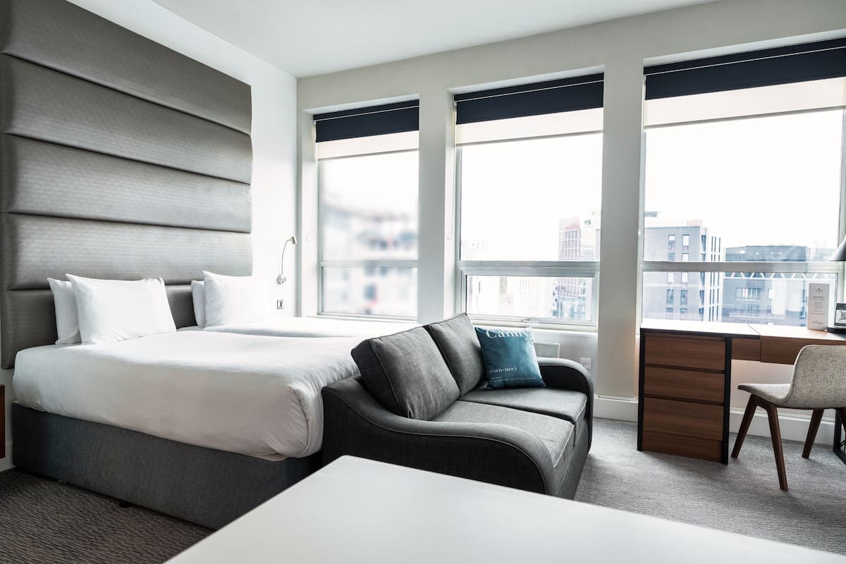 A modern hotel room with a large bed, a sofa, a desk, and a view of the city through large windows.