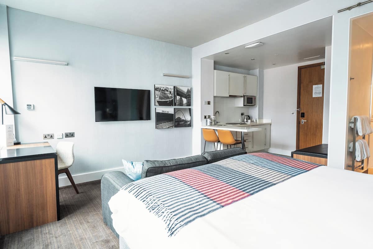 A modern hotel room with a bed, a desk, a chair, a television, and a kitchenette.