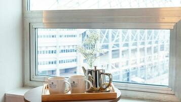 A tray with two white mugs and a silver teapot on a wooden table by a window with a view of a building.
