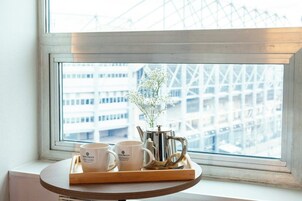 A tray with two white mugs and a silver teapot on a wooden table by a window with a view of a building.