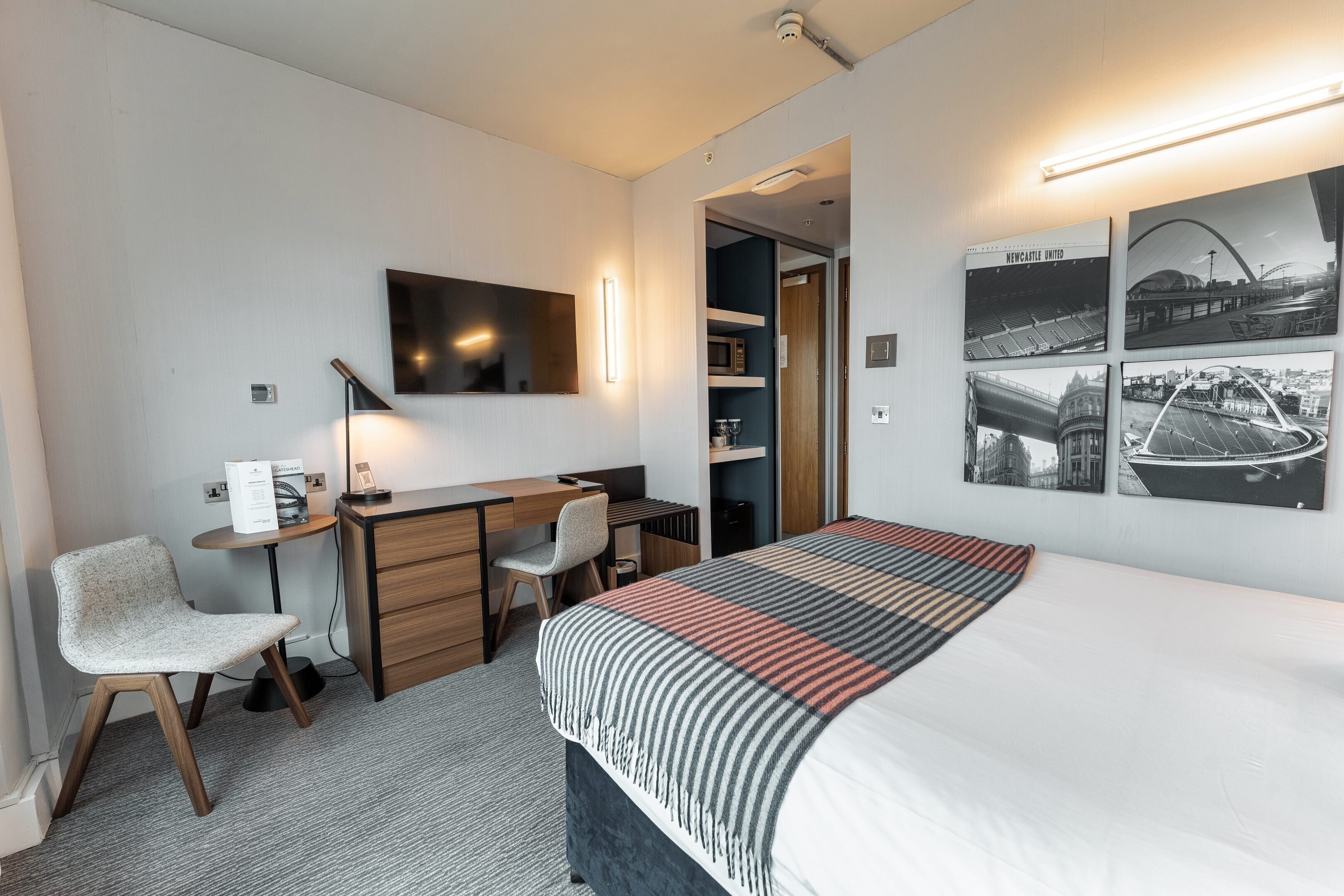 A hotel room with a bed, desk, chair, and a wall-mounted TV.