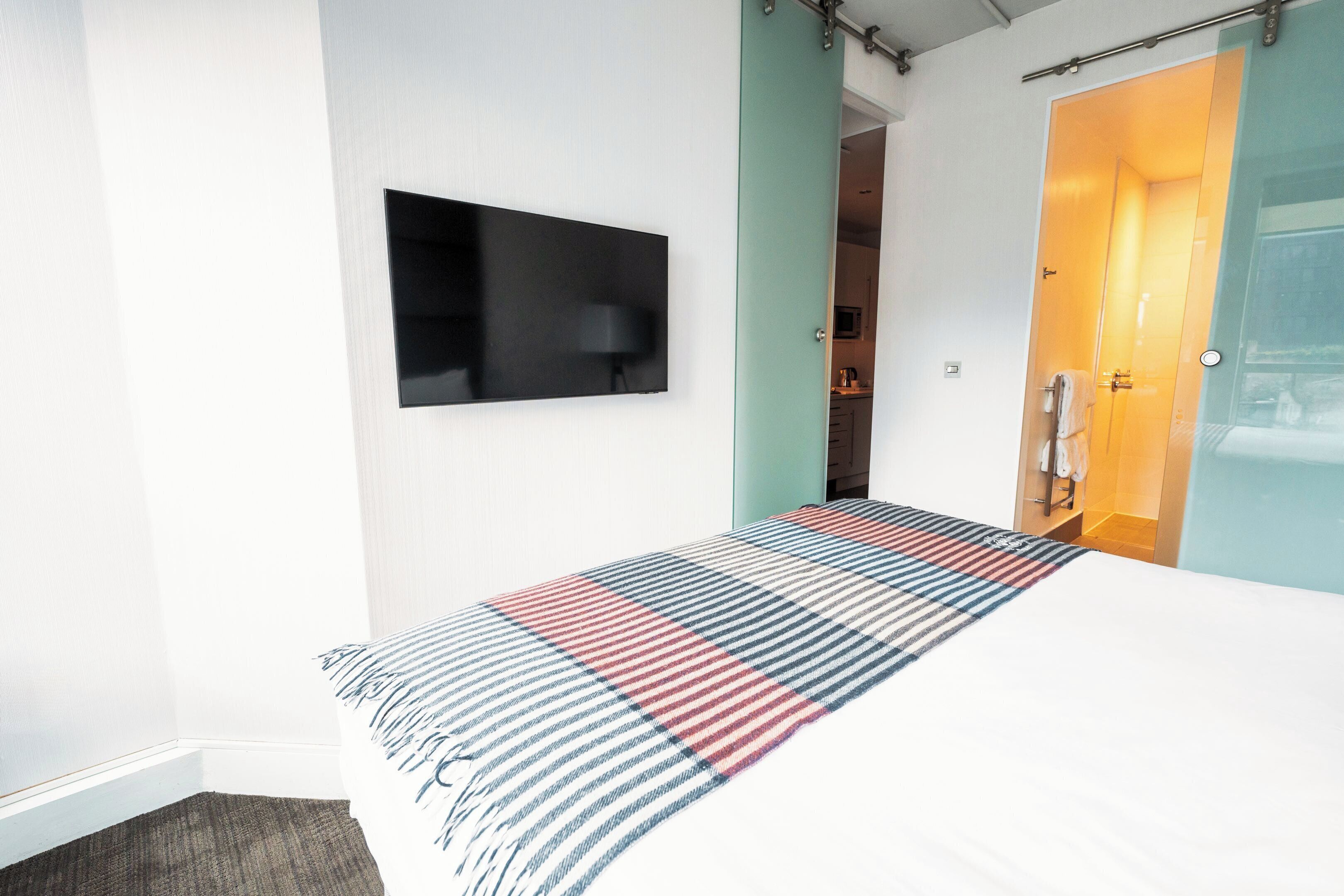 Ett modernt hotellrum med en stor säng, en platt-TV som är monterad på väggen och ett badrum med en dusch med glasdörr.