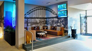A modern lobby with a seating area featuring a sofa and a coffee table, a large screen displaying content, and a cityscape mural on the wall.
