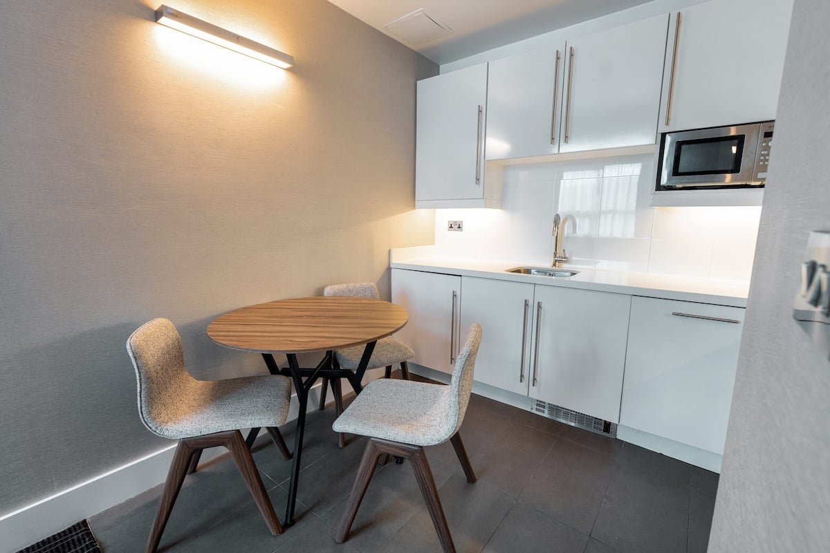 A modern kitchen with white cabinets, a stainless steel microwave, a round wooden table, and two upholstered chairs.