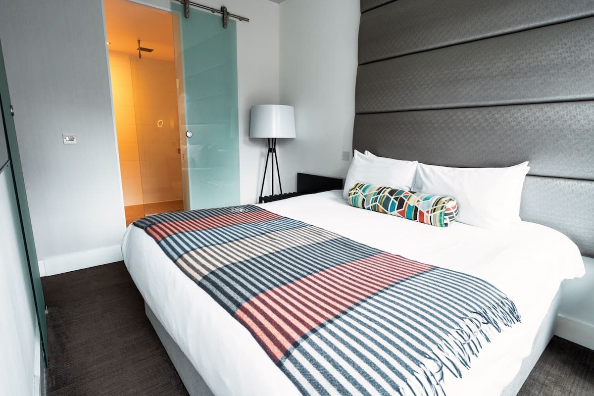 A modern hotel room with a large bed, a glass shower door, a standing lamp, and a grey headboard.