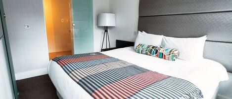 A modern hotel room with a large bed, a glass shower door, a standing lamp, and a grey headboard.