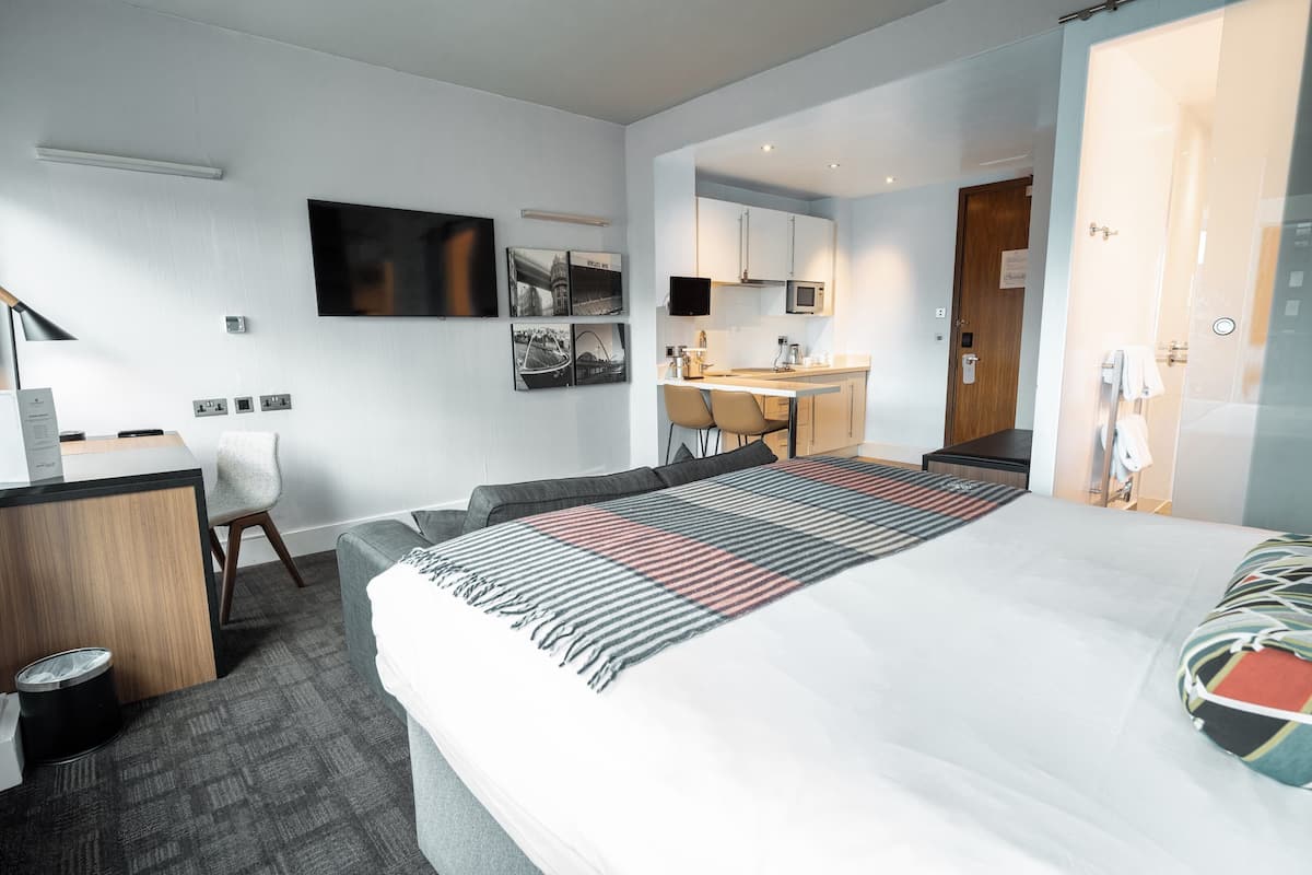 A modern hotel room with a bed, desk, and kitchenette.