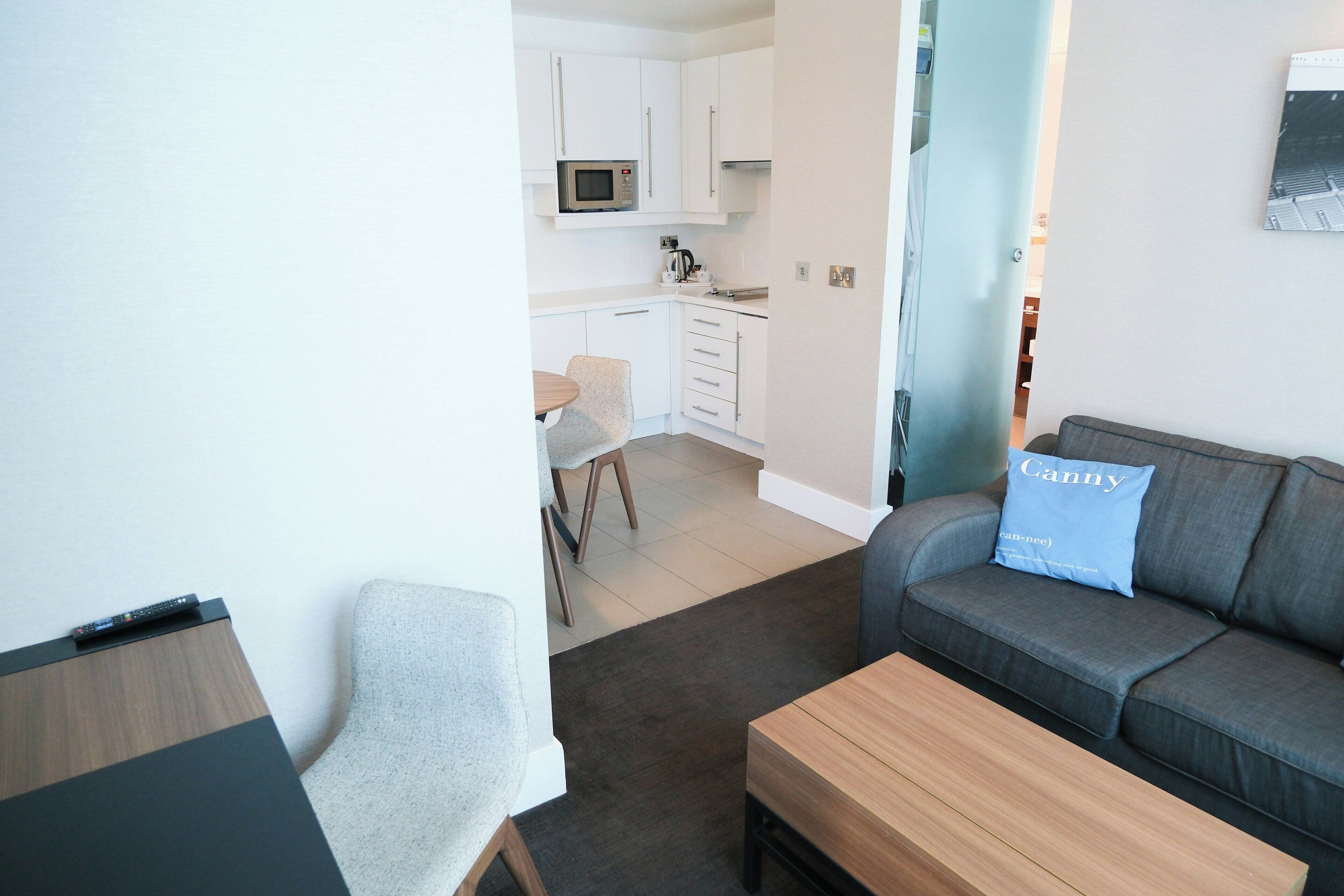 Una sala moderna con un sofá gris, una mesa de centro de madera y un sillón gris. Se ve una cocina con armarios blancos y un microondas al fondo.