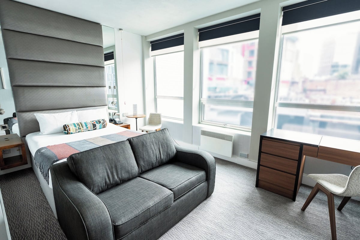 A modern hotel room with a large bed, grey sofa, desk, and chair.