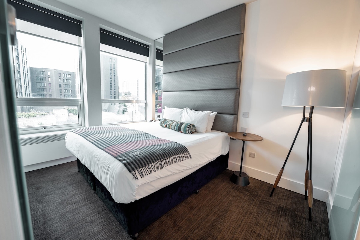 Une chambre d’hôtel moderne, dotée d’un grand lit, d’une table de chevet, d’une lampe et offrant une vue sur la ville grâce à de grandes fenêtres.