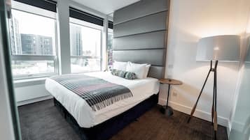 Un moderno cuarto de hotel con una cama grande, una mesita de noche, una lámpara y vistas de la ciudad a través de amplias ventanas.