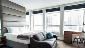 A modern hotel room with a large bed, a sofa, a desk, and large windows offering city views.
