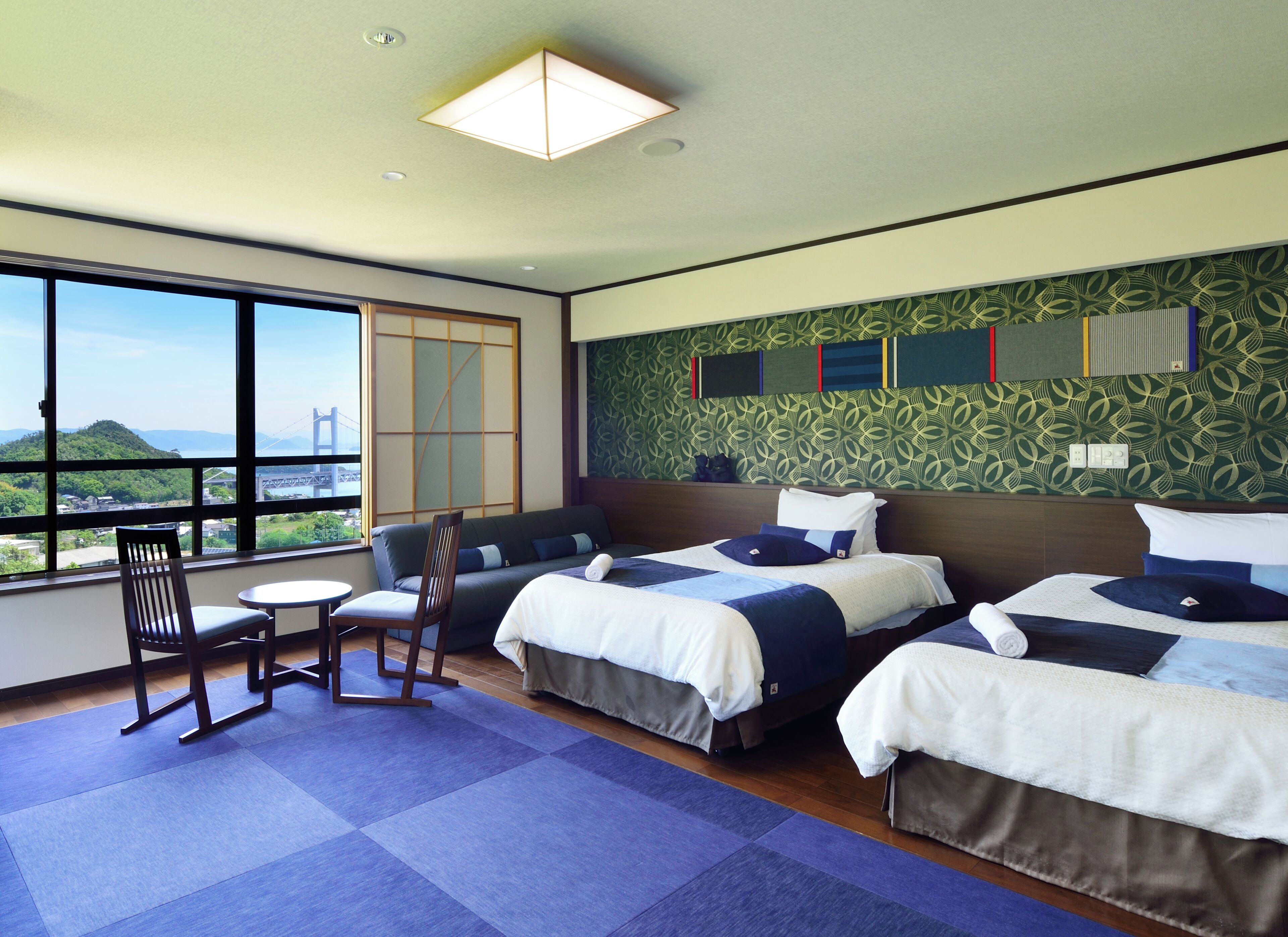A hotel room with two beds, a sofa, a chair, a small table, and a large window offering a view of a bridge and greenery.