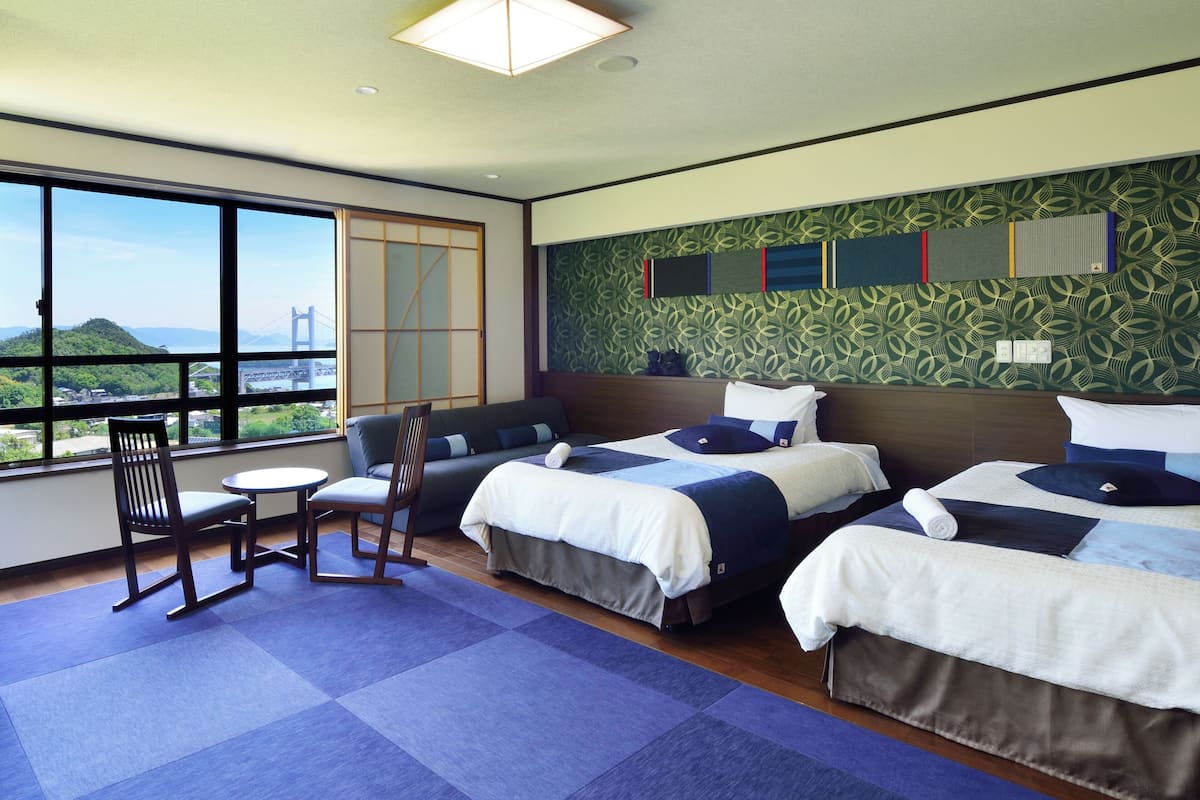 A hotel room with two beds, a sofa, a chair, a small table, and a large window offering a view of a bridge and greenery.