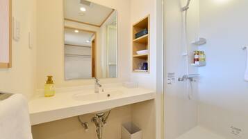 A bathroom with a white sink, mirror, and shower. There are shelves with toiletries and a trash bin.