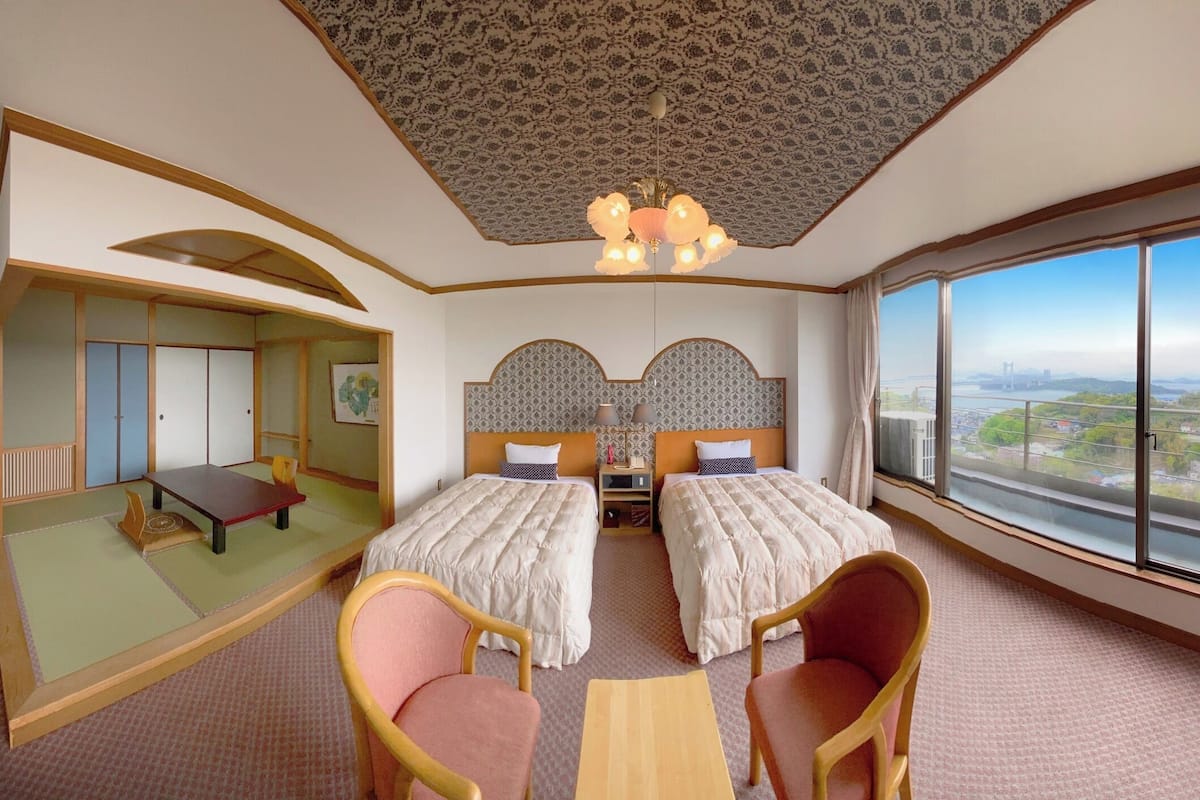 A hotel room with two beds, a sitting area with chairs, a view of a cityscape, and a patterned ceiling.