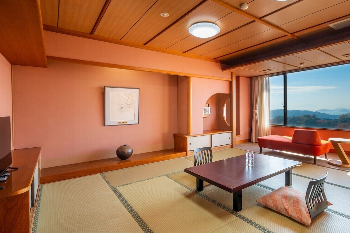 A modern room with tatami flooring, a wooden desk, a sofa, and a large window offering a scenic view.