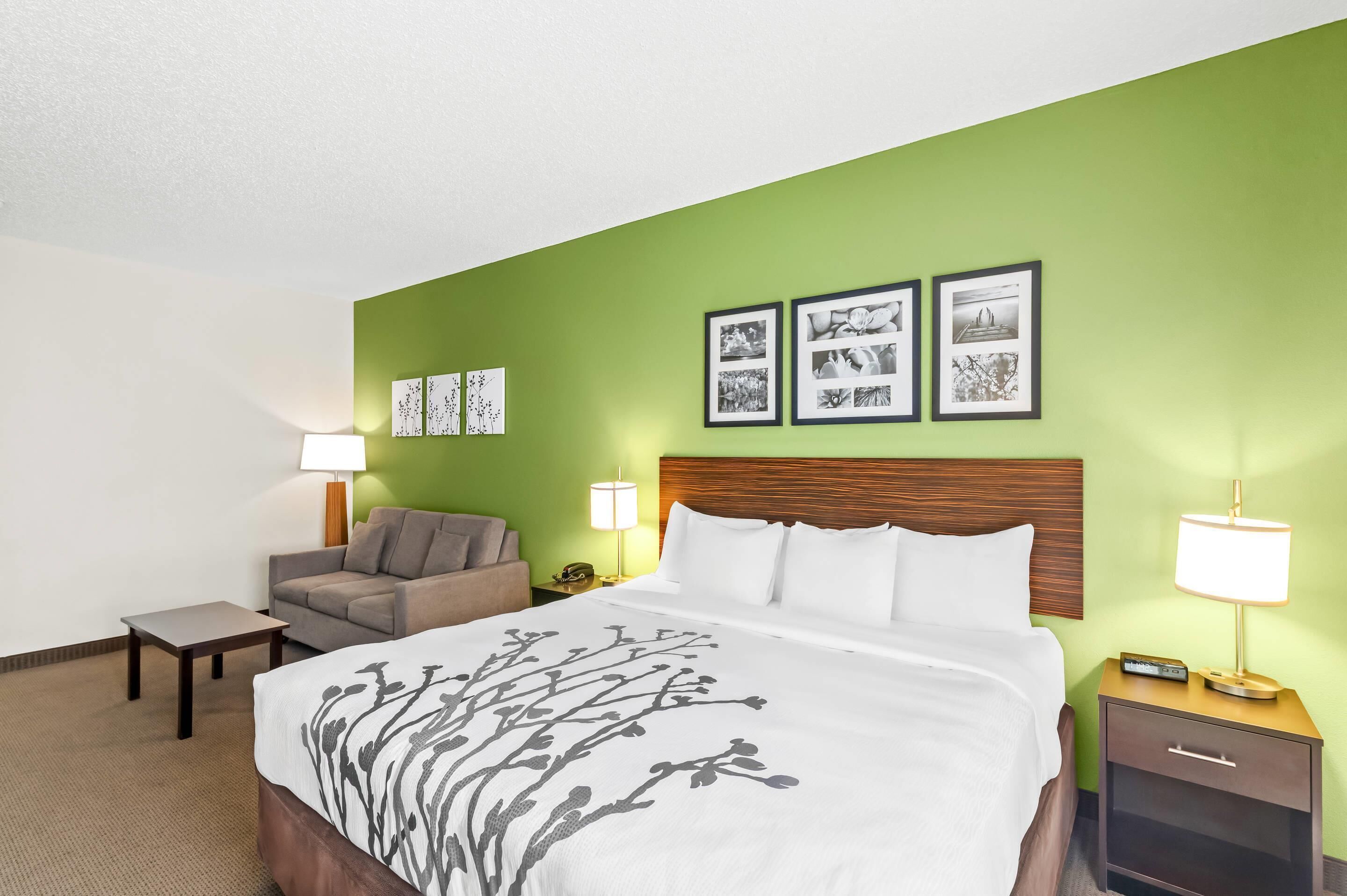 A hotel room with a bed, a nightstand, a lamp, a chair, and a wall with framed pictures.