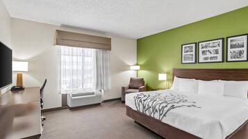 A hotel room with a large bed, a desk with a lamp, a chair, a window with curtains, and a wall with framed pictures.