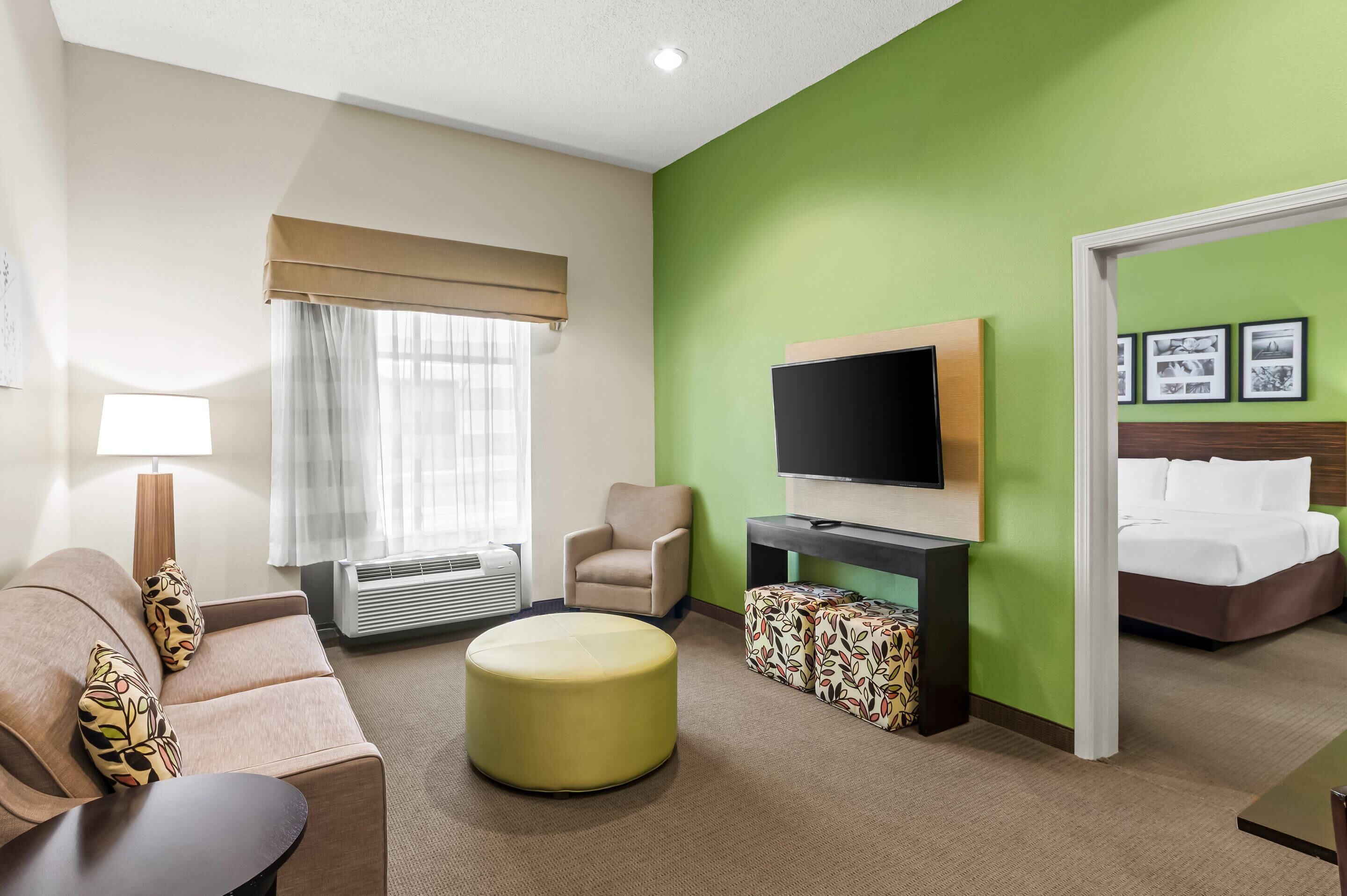 A hotel room with a sofa, ottoman, TV, and a bed.