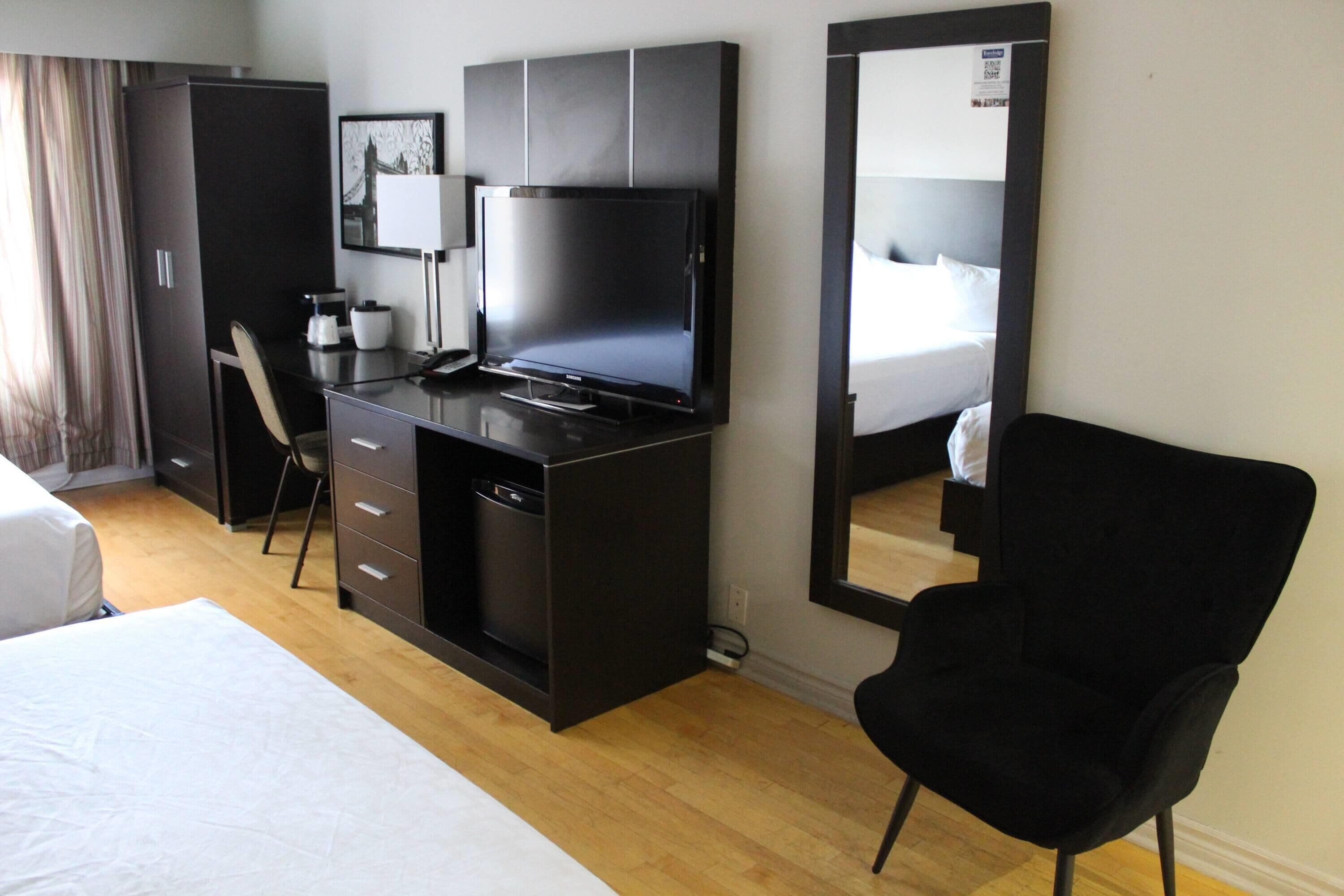 A hotel room with a bed, a desk with a TV, a chair, a wardrobe, and a mirror.