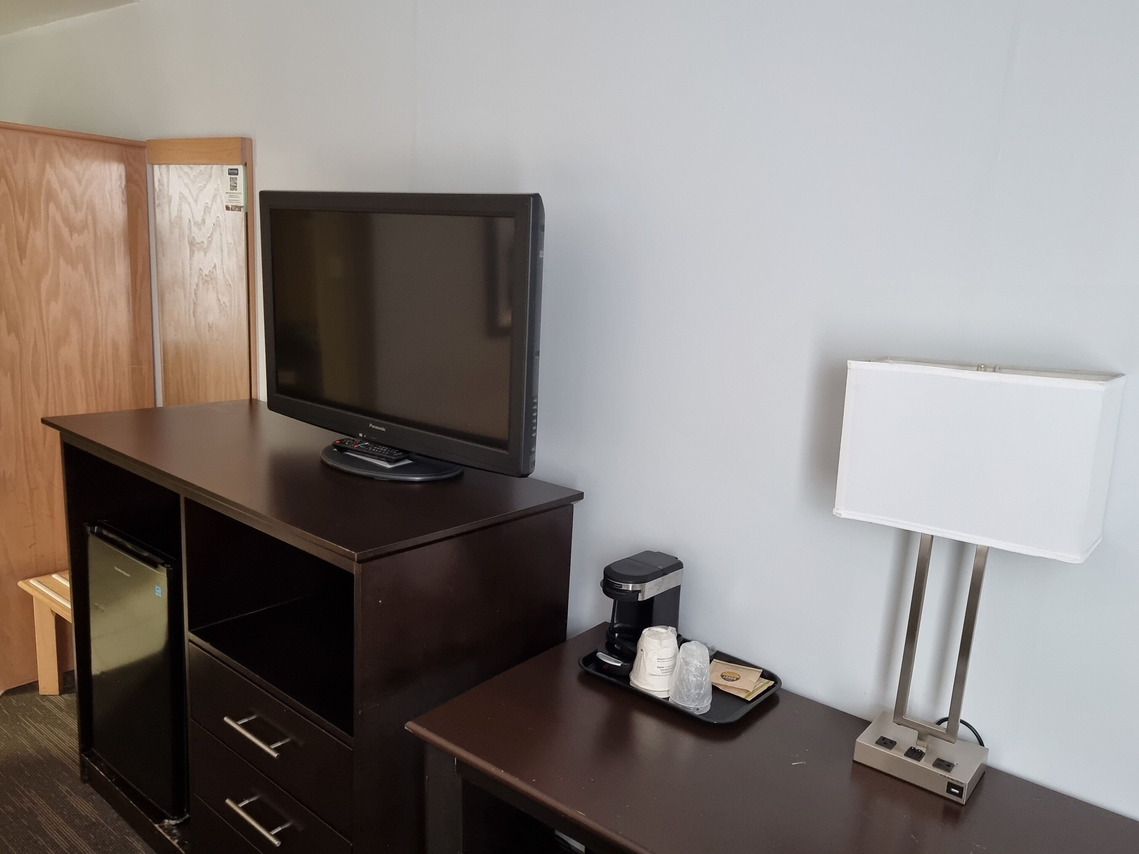 Habitación de hotel con televisión de pantalla plana sobre un escritorio de madera, cafetera, bandeja con tazas y un caja de pañuelos, y una lámpara blanca.